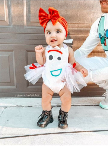 Baby in a forky costume with a red bow, standing on a wooden floor.
