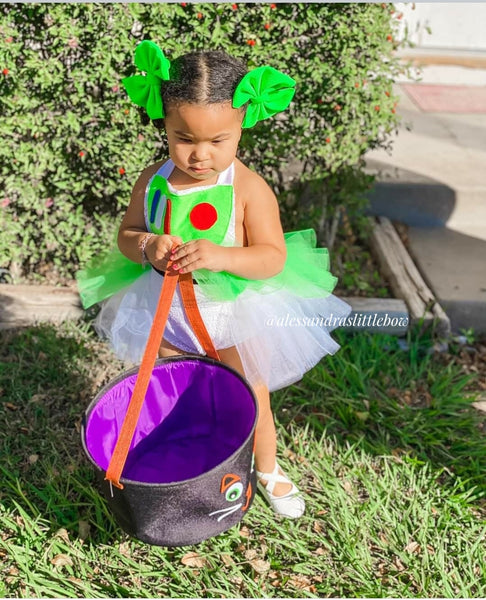 Child in a green and white costume with a purple Halloween bucket outdoors.